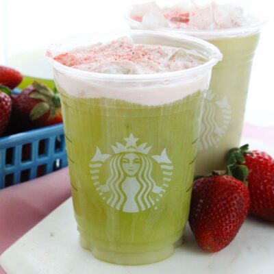 Two Starbucks cups filled with strawberry matcha latte, topped with whipped cream and pink powder, are placed next to fresh strawberries.