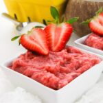 A square white bowl filled with strawberry mousse, made using a delightful strawberry pudding recipe, is topped with two halved fresh strawberries. A yellow container and a spoon are blurred in the background.