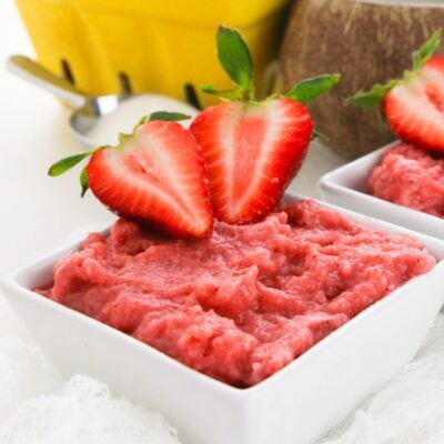 A square white bowl filled with strawberry mousse, made using a delightful strawberry pudding recipe, is topped with two halved fresh strawberries. A yellow container and a spoon are blurred in the background.
