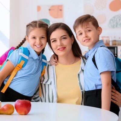 A woman sits at a table with two smiling school-aged children wearing blue uniforms and backpacks. Apples are on the table in front of them.