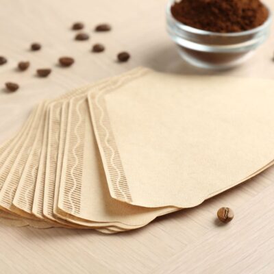 Several brown paper coffee filters are spread out on a light wooden surface, with scattered coffee beans and a small bowl of ground coffee nearby.