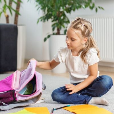 A young girl sitting on the floor opens a pink backpack, with notebooks and stationery spread out in front of her, beginning her after school routine.