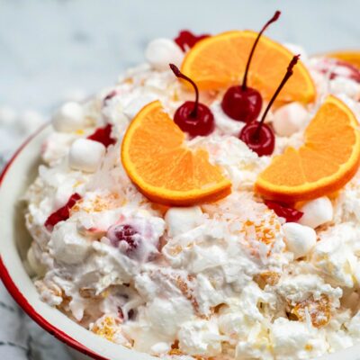 A bowl of ambrosia salad with Cool Whip, orange slices, maraschino cherries, mini marshmallows, and shredded coconut.