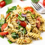 A plate of rotini pasta salad with spinach, cherry tomatoes, mozzarella, grilled chicken, pine nuts, and shredded cheese, garnished with pesto sauce.
