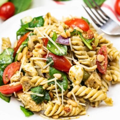 A plate of rotini pasta salad with spinach, cherry tomatoes, mozzarella, grilled chicken, pine nuts, and shredded cheese, garnished with pesto sauce.