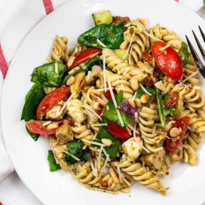 A white plate of rotini pasta salad with spinach, cherry tomatoes, mozzarella balls, pine nuts, and shredded cheese, set on a red-striped towel with a fork beside it.