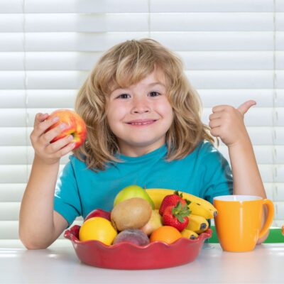 A smiling child with long blond hair holds an apple and gives a thumbs up, sitting at a table with tasty after school snacks—a bowl of assorted fruit and an orange mug.