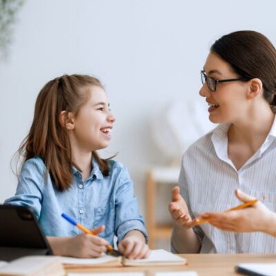 An adult and a child sit at a table, smiling and talking while writing in notebooks, with a tablet and stationery nearby.