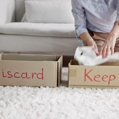 A person sorts items into two cardboard boxes labeled "Discard" and "Keep" on a carpeted floor in front of a sofa.