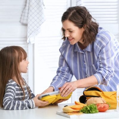 A woman helps with school lunch packing, handing a lunchbox to a young girl at the kitchen counter where a sandwich, vegetables, and fruit are laid out.