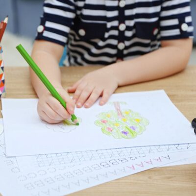 A child in a striped shirt colors a picture of a brain with a green pencil, with kids school work and alphabet practice sheets spread across the desk.