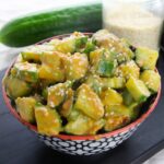 A bowl of cucumber salad with creamy dressing and sesame seeds, with a whole cucumber and a jar of sesame seeds in the background.