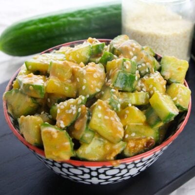 A bowl of cucumber salad with creamy dressing and sesame seeds, with a whole cucumber and a jar of sesame seeds in the background.