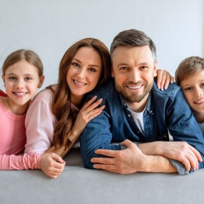 A family of four, two adults and two children, sit closely together on a couch, smiling at the camera in a bright living room.