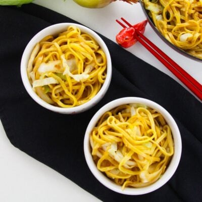 Two white bowls filled with chow mein with sauce and cabbage are placed on a black cloth, with red chopsticks and a decorative figurine nearby.