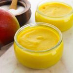 Two small glass jars filled with smooth, yellow mango mousse are placed on a white surface, alongside a whole mango and a wooden container of salt—perfect for fans of creamy mango pudding.