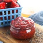 A small glass jar filled with strawberry chia seed jam sits on a wooden surface next to scattered chia seeds and a blue basket of fresh strawberries.