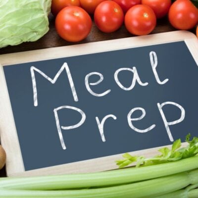 A chalkboard with "Meal Prep" written on it is surrounded by fresh vegetables including potatoes, celery, carrots, cabbage, tomatoes, and cucumber.
