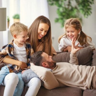 A man lies on a sofa talking to a woman, young boy, and young girl—all smiling and gathered closely together in a bright living room, sharing family bonding time with a question a day.