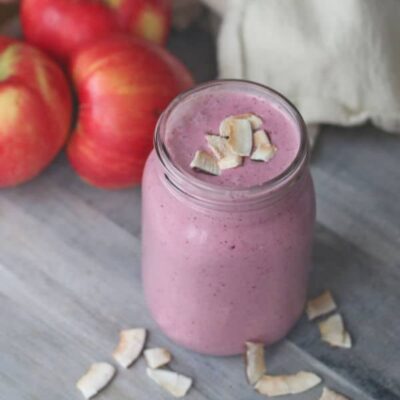 A jar of pink smoothie topped with coconut flakes sits on a wooden surface, with red apples and a few coconut flakes beside it.