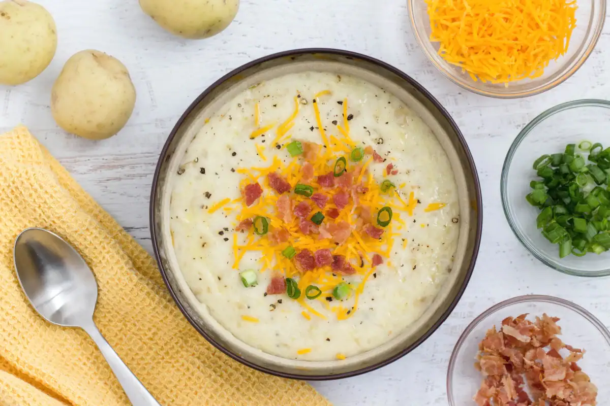 A bowl of potato soup topped with shredded cheese, bacon bits, and chopped green onions, next to the ingredients in bowls.
