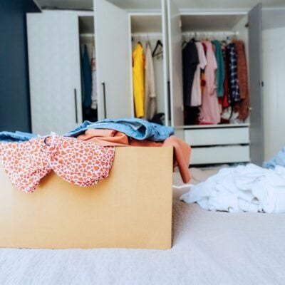 A cardboard box filled with clothes sits on a bed, with more clothes scattered around and an open closet visible in the background.