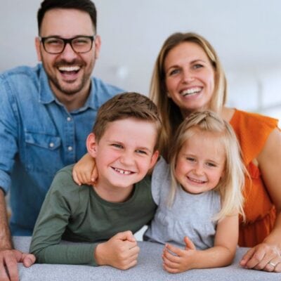 A man, a woman, a young boy, and a young girl are smiling and posing together indoors, leaning over the back of a couch.