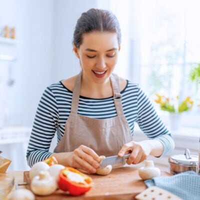 A woman wearing an apron slices mushrooms on a wooden cutting board in a bright kitchen surrounded by various vegetables and cooking utensils.