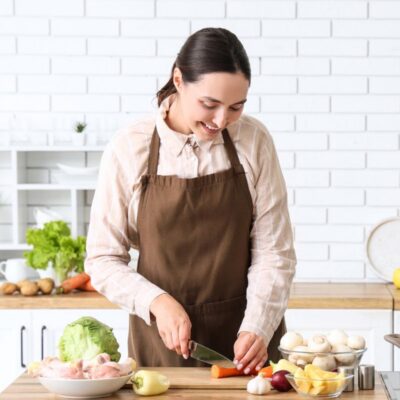 Woman wearing a brown apron slices vegetables on a wooden counter in a modern kitchen with various fresh ingredients around her.