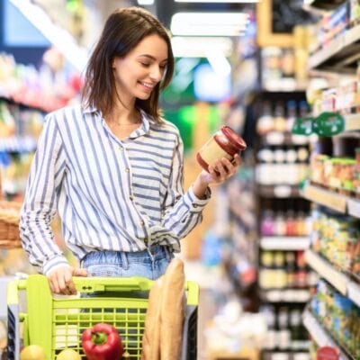 A woman stands in a grocery store aisle, holding a jar and examining its label, with a shopping cart containing groceries such as bread and produce.