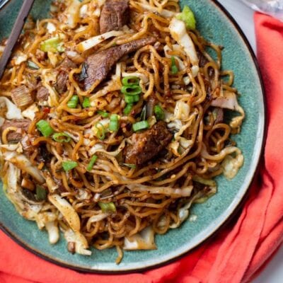 A plate of stir-fried noodles with beef, cabbage, and green onions, served with chopsticks on the side and a red napkin underneath.
