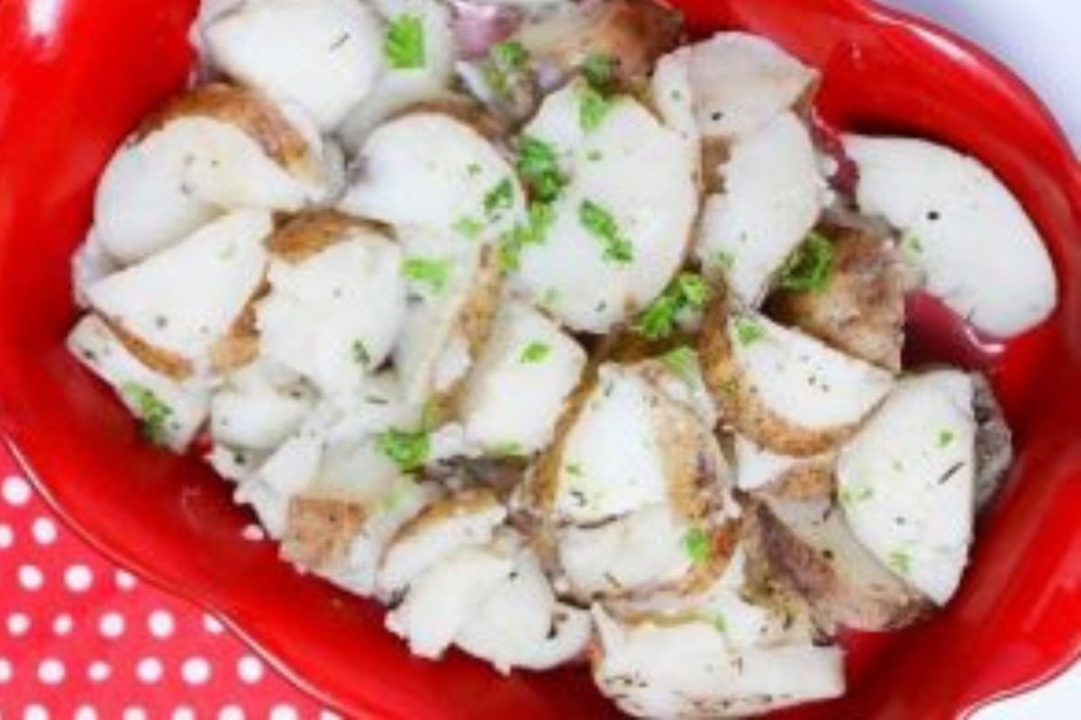 A red dish filled with sliced potatoes garnished with chopped parsley, placed on a polka dot cloth.