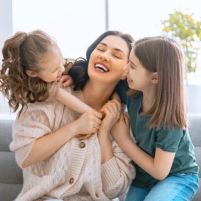 A woman sits on a couch smiling and laughing with two young girls, one on each side of her, in a bright living room.