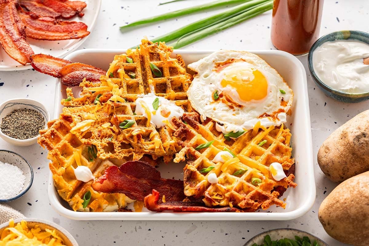 Waffles on a plate topped with sour cream, green onions, bacon and a sunny-side-up egg next to bowls of the ingredients.