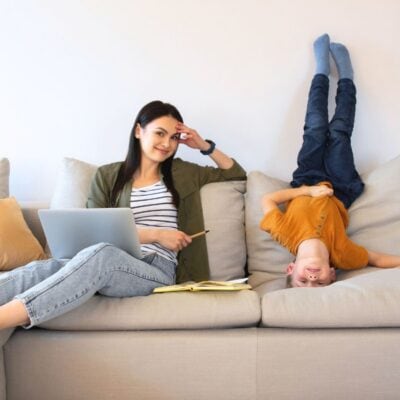A woman sits on a couch with a laptop and notebook while a child lies upside down beside her, legs up against the couch back.
