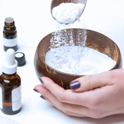 A person pours white powder into a wooden bowl beside small glass dropper bottles on a white surface.