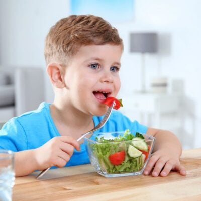 A young boy in a blue shirt sits at a table and eats a salad with a fork.