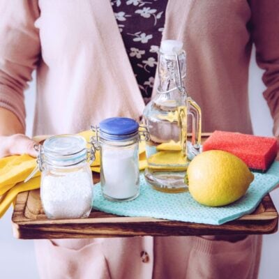 A person holds a tray with cleaning items: gloves, baking soda, salt, a jar of vinegar, a lemon, sponges, and cloths.