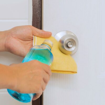 A person pours cleaning liquid onto a yellow cloth while cleaning a silver door knob on a white door.
