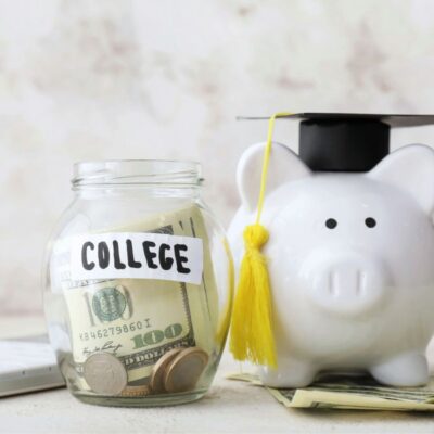 A piggy bank with a graduation cap sits next to a jar labeled "College" filled with cash, representing saving money for education.