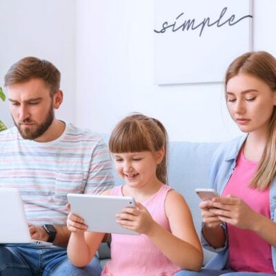 A man, a young girl, and a woman sit on a couch using a laptop, tablet, and smartphone respectively, with a plant and a framed "simple" sign in the background.