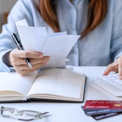 A person reviews receipts, writes in a notebook, and uses a calculator at a desk with credit cards, glasses, and a smartphone nearby.