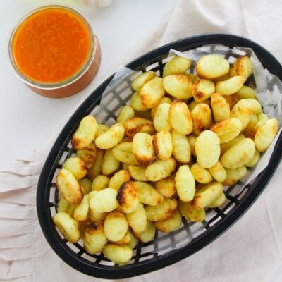 A basket of golden-brown crispy gnocchi sits on parchment paper next to a glass container of red dipping sauce.