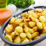 A basket of crispy baked gnocchi with char marks, served with a side of red dipping sauce. Fresh herbs and a garlic bulb are blurred in the background.