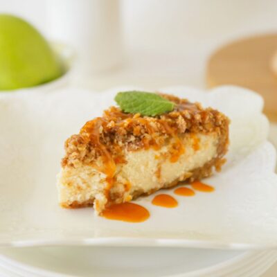 A slice of cheesecake topped with crumble, caramel sauce, and a mint leaf sits on a white plate.