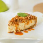 A slice of cheesecake with a crumbly topping, garnished with a mint leaf and drizzled with caramel sauce, served on a white plate.