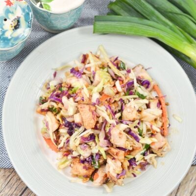 A plate of mixed salad with shredded cabbage, carrots, green onions, and pieces of grilled chicken, with additional green onions and a bowl of sauce nearby.