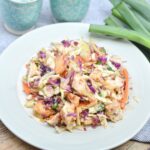A plate of Asian-style chicken salad with shredded cabbage, carrots, and purple cabbage, placed on a white plate next to green onions and patterned cups.