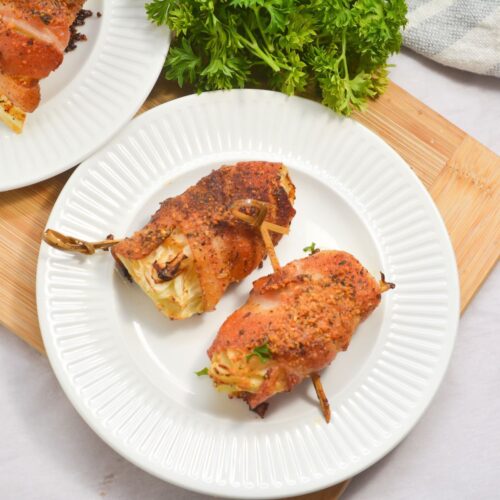 Two pieces of seasoned, cooked chicken breast wrapped around a filling and secured with toothpicks, served on a white plate beside a bunch of fresh parsley.