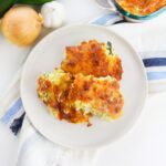 A white plate with two pieces of baked zucchini casserole, placed on a striped cloth next to an onion, garlic, and part of a casserole dish.